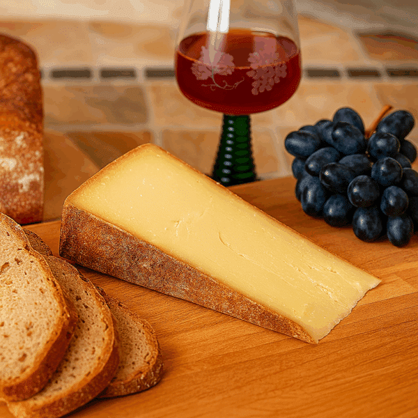 Ein Stück 2-3 Jahre alter Bergkäse aus dem Bregenzerwald auf einem Holzbrett, serviert mit Bauernbrot, blauen Trauben und einem Glas Rotwein.