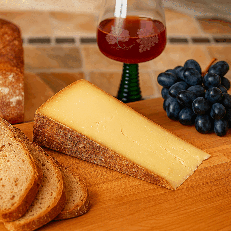 Ein Stück 2-3 Jahre alter Bergkäse aus dem Bregenzerwald auf einem Holzbrett, serviert mit Bauernbrot, blauen Trauben und einem Glas Rotwein.