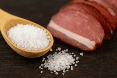 Körniger Salz und luftgetrocknetes Fleisch - ALMGOLD Grobkörniges Nitritpökelsalz in einem Holzlöffel auf dunklem Untergrund – Symbolbild für Konservierung, Fleischverarbeitung und Lebensmittelhygiene